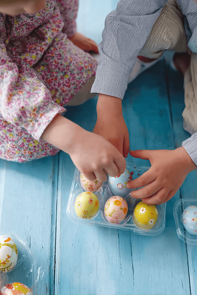 two children sitting on a bright pastel blue wooden floor, reaching into a clear plastic egg carton filled with decorated Easter eggs.
