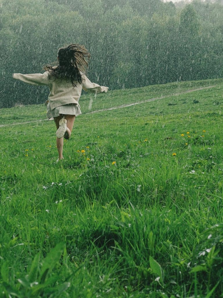 a carefree young woman running barefoot through a lush, green meadow in the rain.
