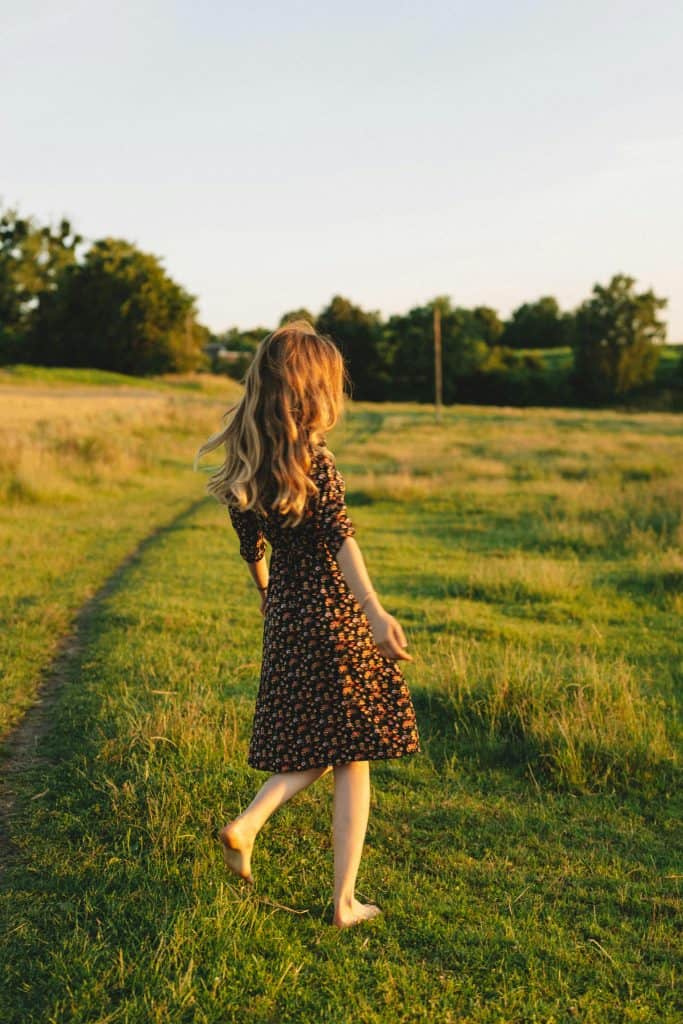 a young girl walking barefoot through a sunlit countryside field during golden hour.