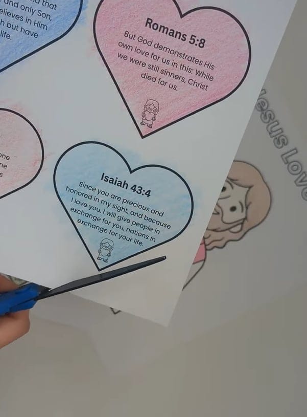 A hand holding blue scissors and cutting out a blue heart shape with printed text, alongside a pink heart with printed text on a white sheet of paper with a partially visible coloring page underneath