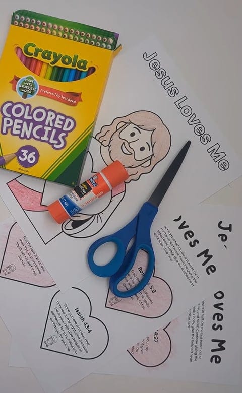 A Crayola 36-count colored pencils box and an orange Elmer's glue stick resting on printed 'Jesus Loves Me' coloring sheets spread out on a white surface, with a blue-handled pair of scissors on top of the papers