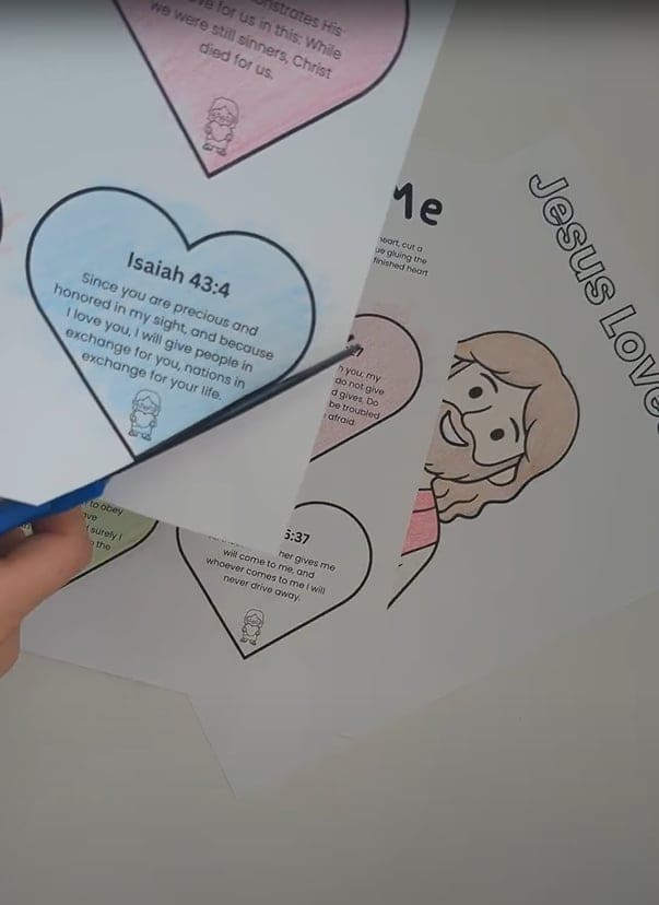 A child's hand holding scissors and cutting out a blue heart-shaped paper with printed text, with multiple other cut-out heart shapes and a 'Jesus Loves Me' coloring sheet visible on a white surface