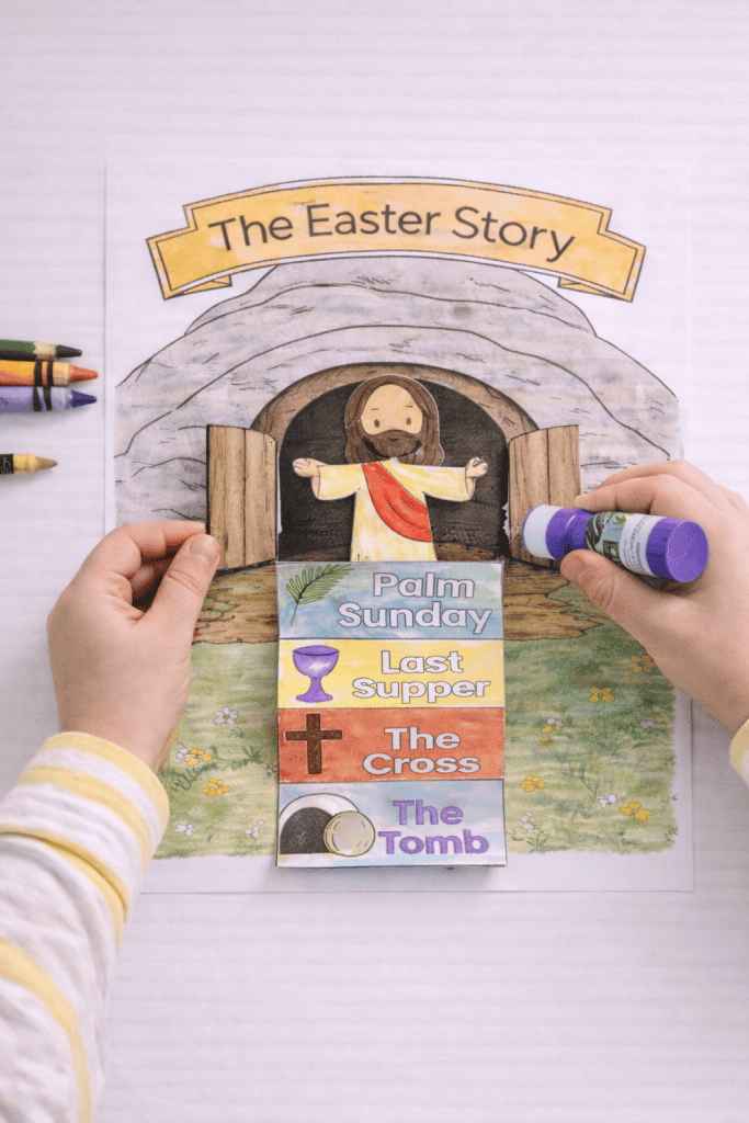 Top-down view of a completed Easter craft on a white table, showing “The Easter Story” tomb page with Jesus inside and two attached tomb doors that open outward