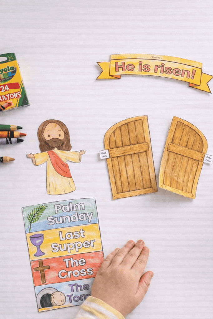 Child’s hand resting on a white table beside freshly cut Easter craft pieces, including a colored Jesus cutout, two wooden tomb doors, a “He is risen!” banner, and a colored story strip, with a Crayola crayon box and crayons nearby.