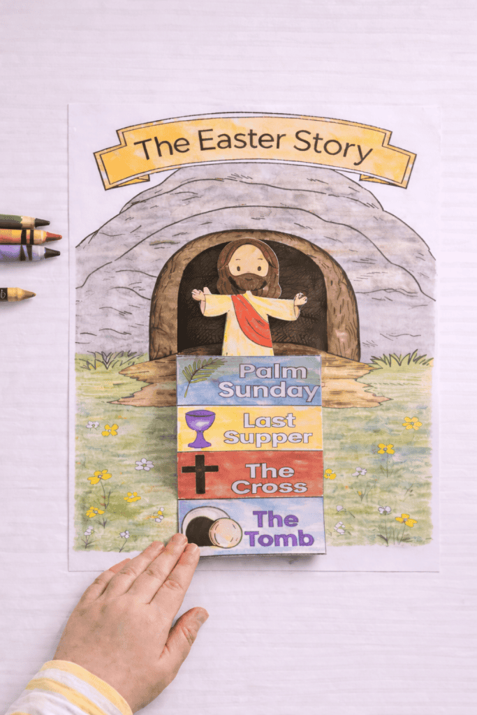 Top-down view of a printed “The Easter Story” tomb page on a white table with a colored Jesus cutout glued inside the tomb opening and a folded story strip attached below
