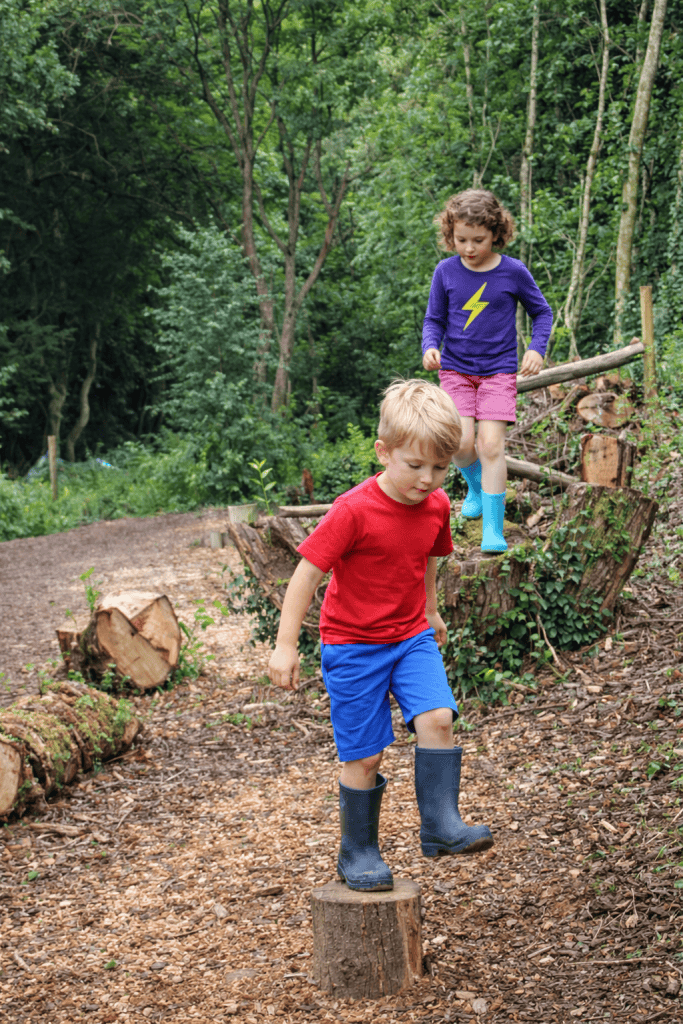 two young children navigating a natural woodland obstacle course made from logs, tree stumps, and uneven forest terrain. 