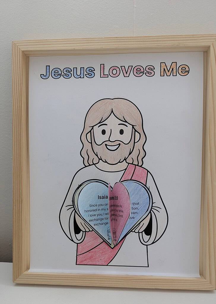 A wooden picture frame standing upright against a white wall, containing a completed 'Jesus Loves Me' colored coloring page showing a cartoon figure with light brown hair holding layered blue and pink paper hearts with printed text