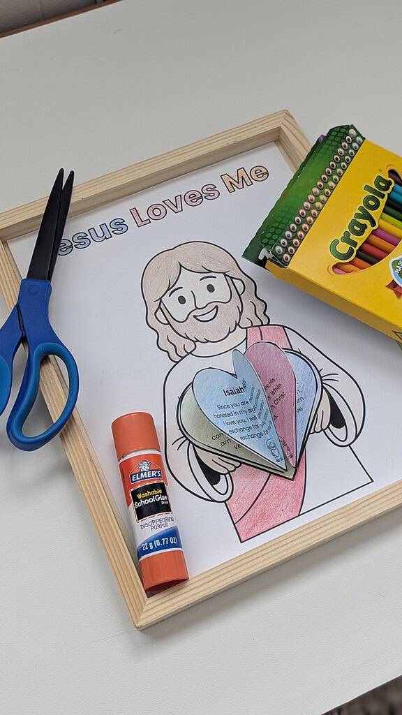 A wooden picture frame with a completed colored 'Jesus Loves Me' coloring page featuring a cartoon figure holding layered paper hearts, displayed on a white surface alongside blue scissors, an Elmer's glue stick, and a Crayola colored pencils box