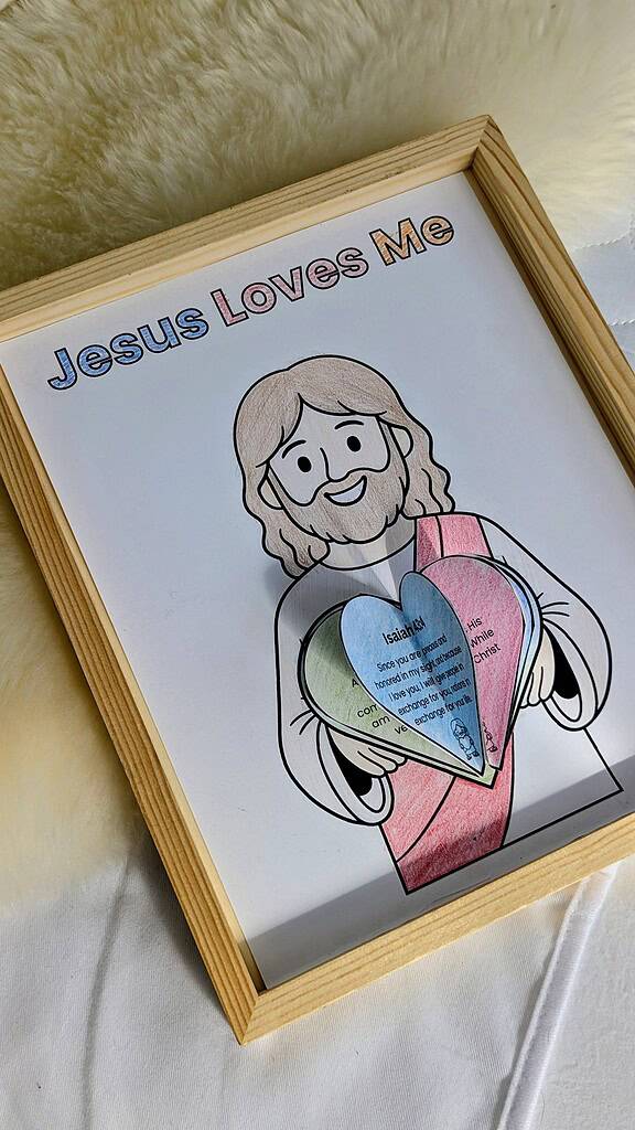 A completed 'Jesus Loves Me' colored artwork inside a wooden picture frame, featuring a cartoon figure with colorful hair holding layered paper hearts with printed text, placed on a white fluffy surface