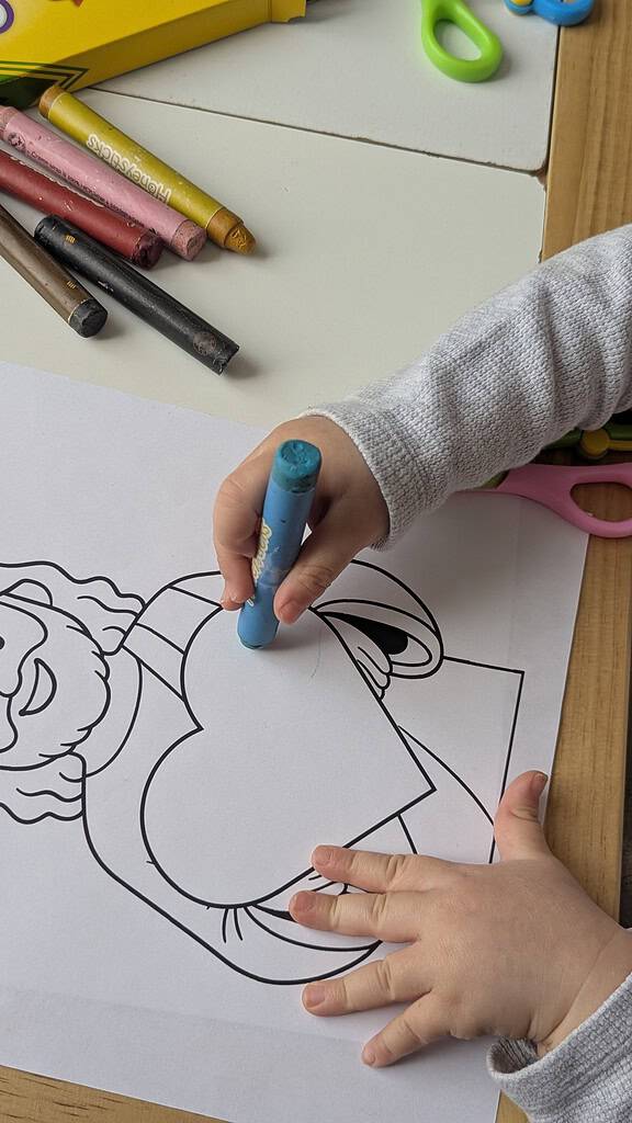 A close-up of two small children's hands, one holding a blue crayon and coloring a black-and-white coloring sheet on a wooden table, with several loose crayons and a pink pair of scissors visible at the edges