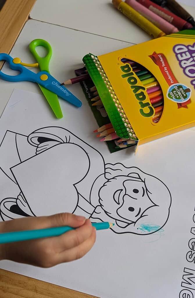 A child's hand holding a teal colored pencil and coloring a cartoon figure on a coloring sheet, with a Crayola colored pencils box and blue-and-green children's scissors visible in the background on a wooden table