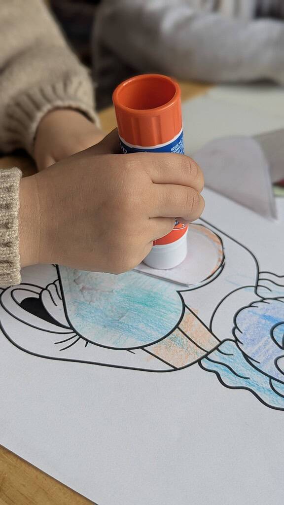 A close-up of a small child's hand pressing an orange-capped glue stick onto a partially colored coloring sheet showing a cartoon figure, on a wooden table