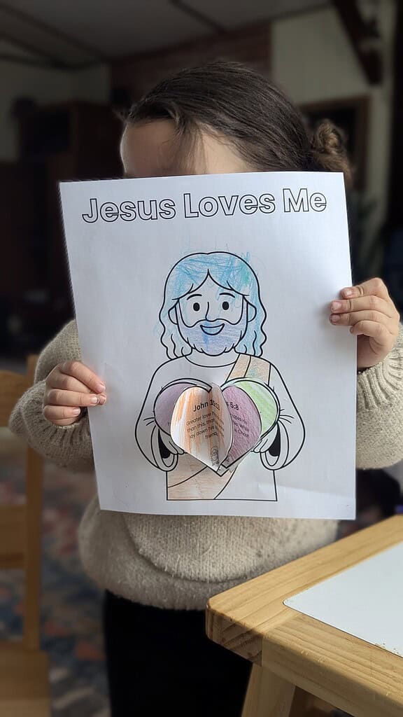 A young child holding up a completed 'Jesus Loves Me' coloring page featuring a colored cartoon figure holding layered paper hearts glued to the page, with the page covering the child's face, in front of a wooden table