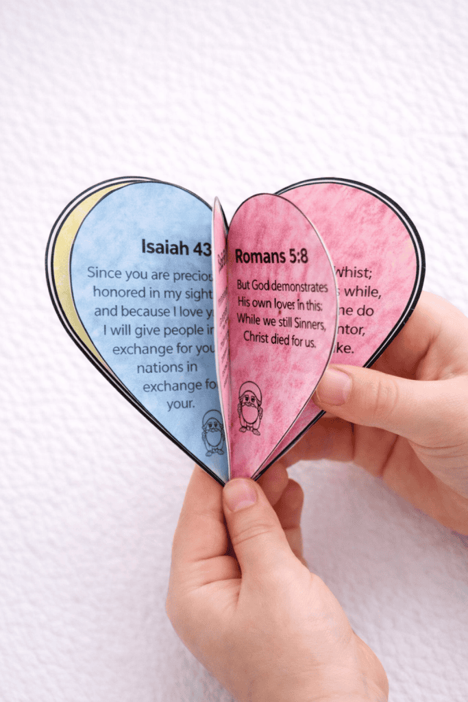 Child holding a folded heart-shaped Christian craft with Bible verses including Isaiah 43:4 and Romans 5:8.