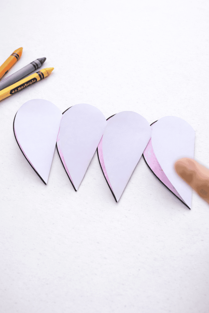 Four folded paper heart halves lined up in a row on a white surface, showing their pink-colored inside edges, with a finger touching the last one and a few crayons visible in the upper left corner