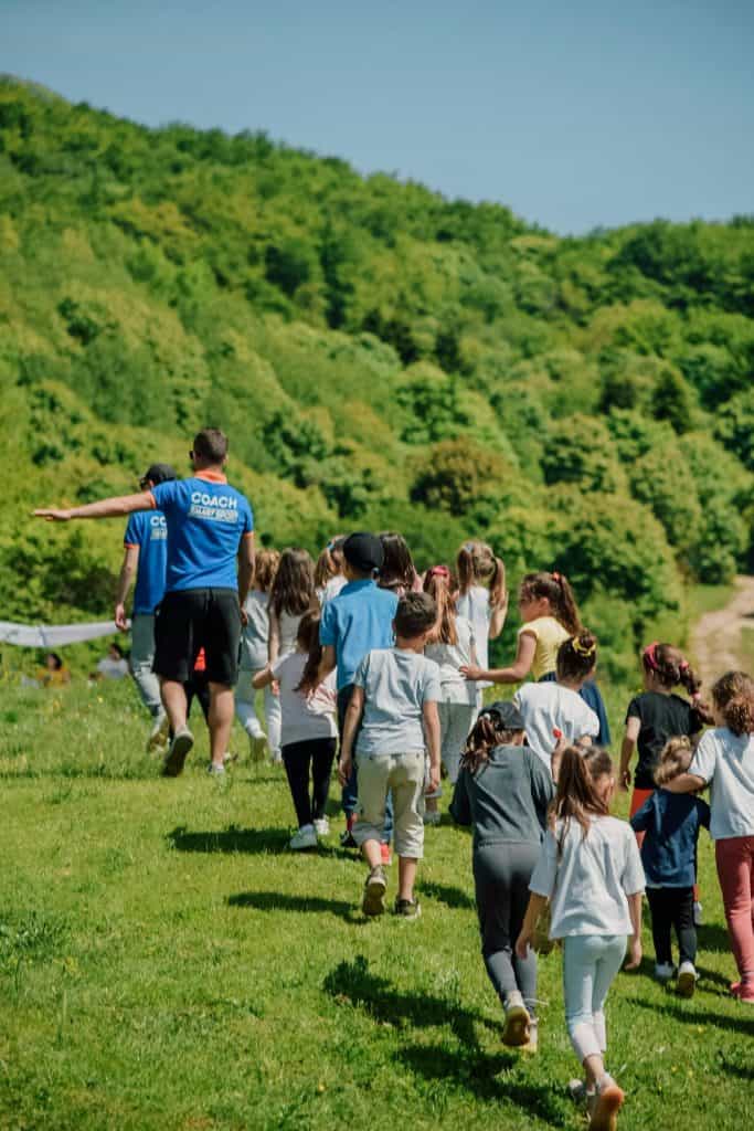 a group of young children walking together across a lush green meadow, guided by an adult coach wearing a bright blue shirt