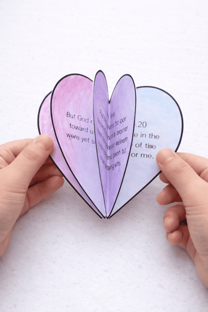 Two hands holding open a layered paper heart craft made of multiple colored heart halves in pink, purple, and blue glued together and fanned out, with printed text visible on each layer, against a white background