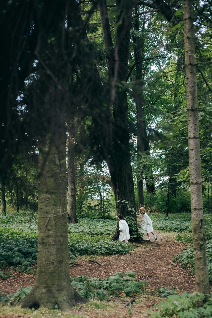 two young children exploring a woodland path, partially hidden among tall trees and dense greenery