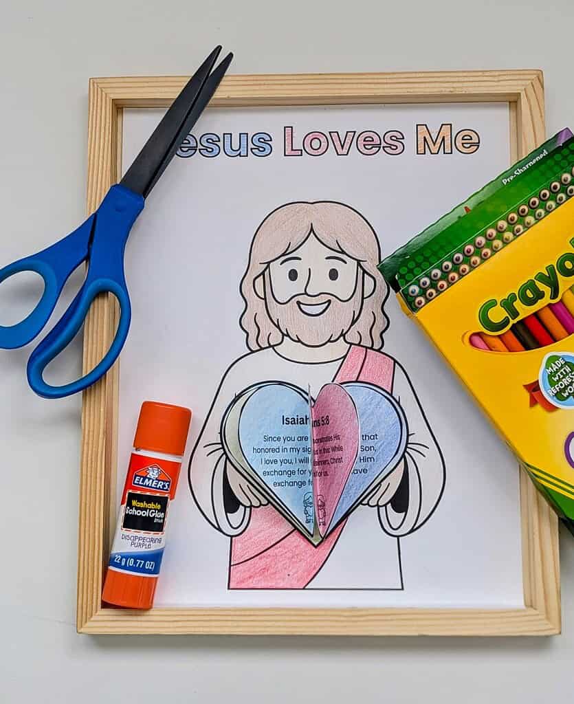 A wooden picture frame displaying a coloring page with 'Jesus Loves Me' title, featuring a cartoon figure holding two colored hearts with text, surrounded by blue scissors, an Elmer's glue stick, and a Crayola colored pencils box on a white surface