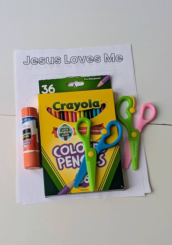 A Crayola 36-count colored pencils box, an Elmer's glue stick, and two pairs of children's scissors in blue-green and pink-green, all laid on top of a printed 'Jesus Loves Me' coloring sheet on a white surface
