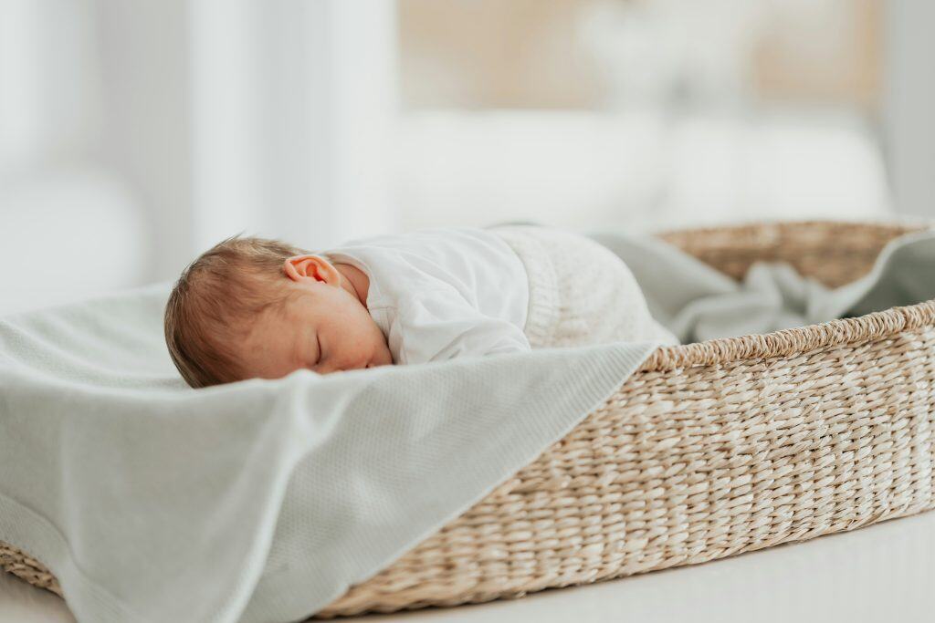 Newborn baby sleeping in a woven bassinet with soft organic cotton bedding in a neutral, minimalist nursery.