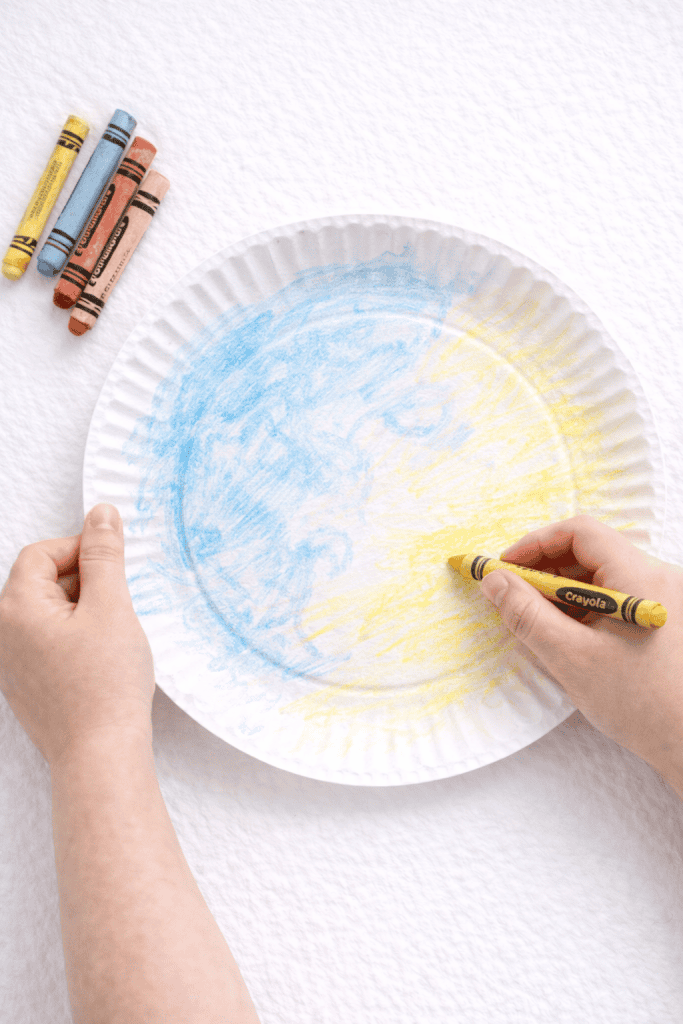 Child coloring a paper plate with blue and yellow crayons
