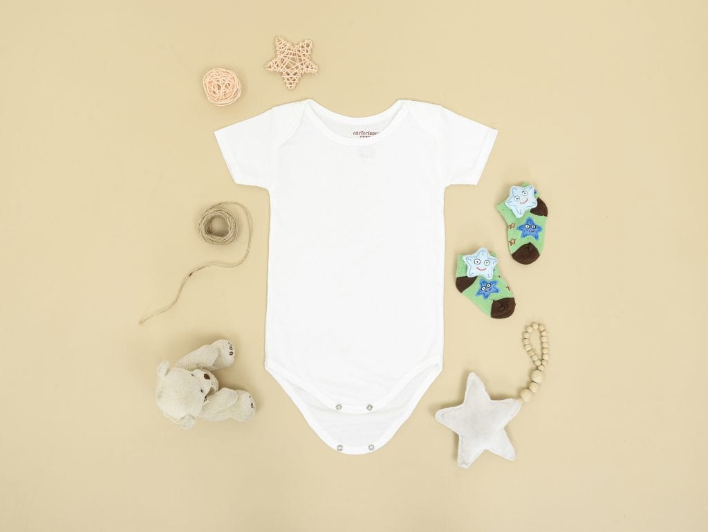 White baby onesie laid flat with socks and soft toys in a neutral, minimalist baby setup.
