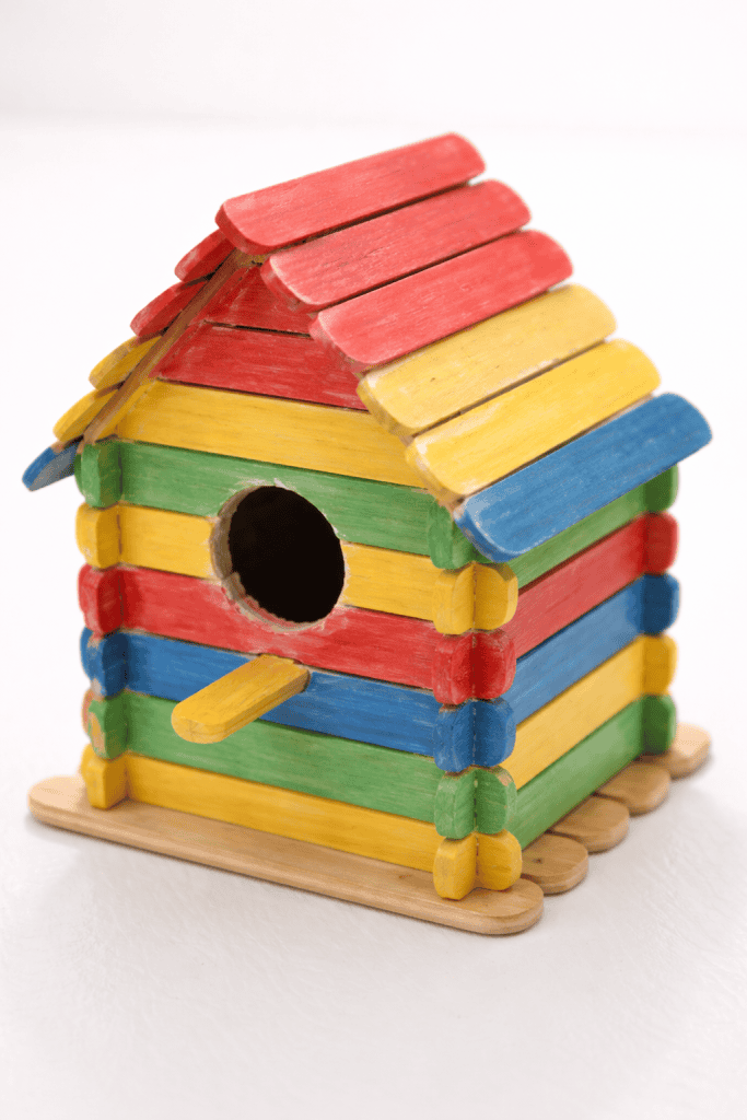 a kid-made popsicle stick birdhouse placed on a plain bright white table.