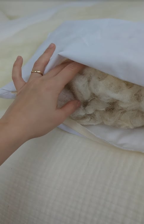 Hand placing natural wool stuffing inside a white pillow cover through the zipper opening.