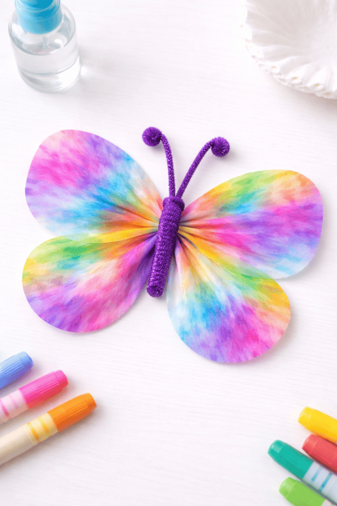 Colorful coffee filter butterfly craft with blended watercolor wings and pipe cleaner body on a white table.