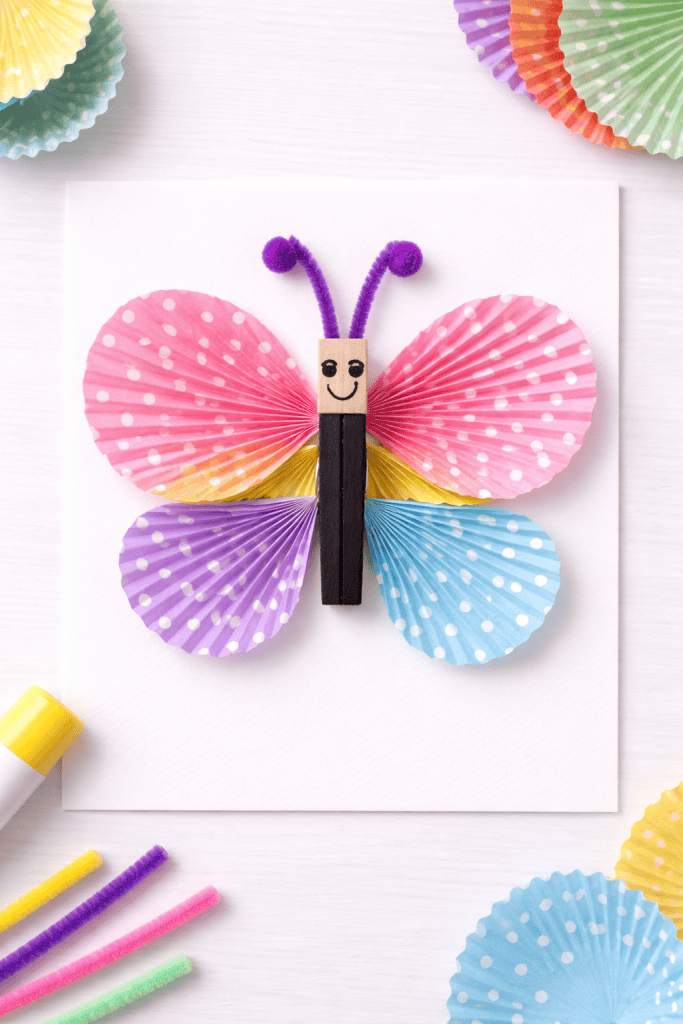 Cupcake liner butterfly craft with colorful wings and clothespin body on white paper.