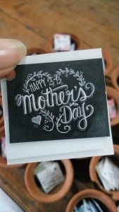 hand holding a happy mother’s day card with decorative lettering and small clay pots in the background