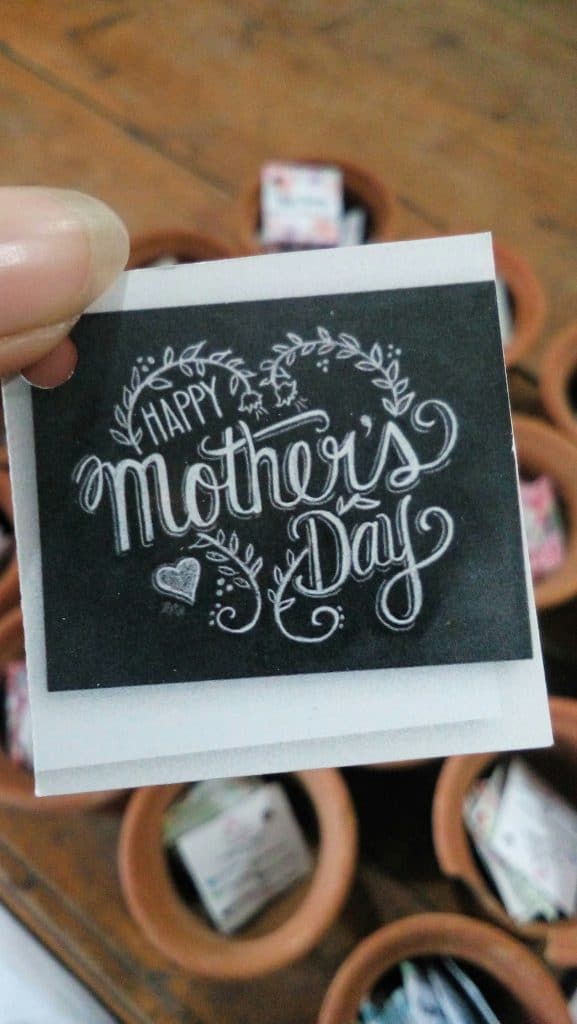 hand holding a happy mother’s day card with decorative lettering and small clay pots in the background