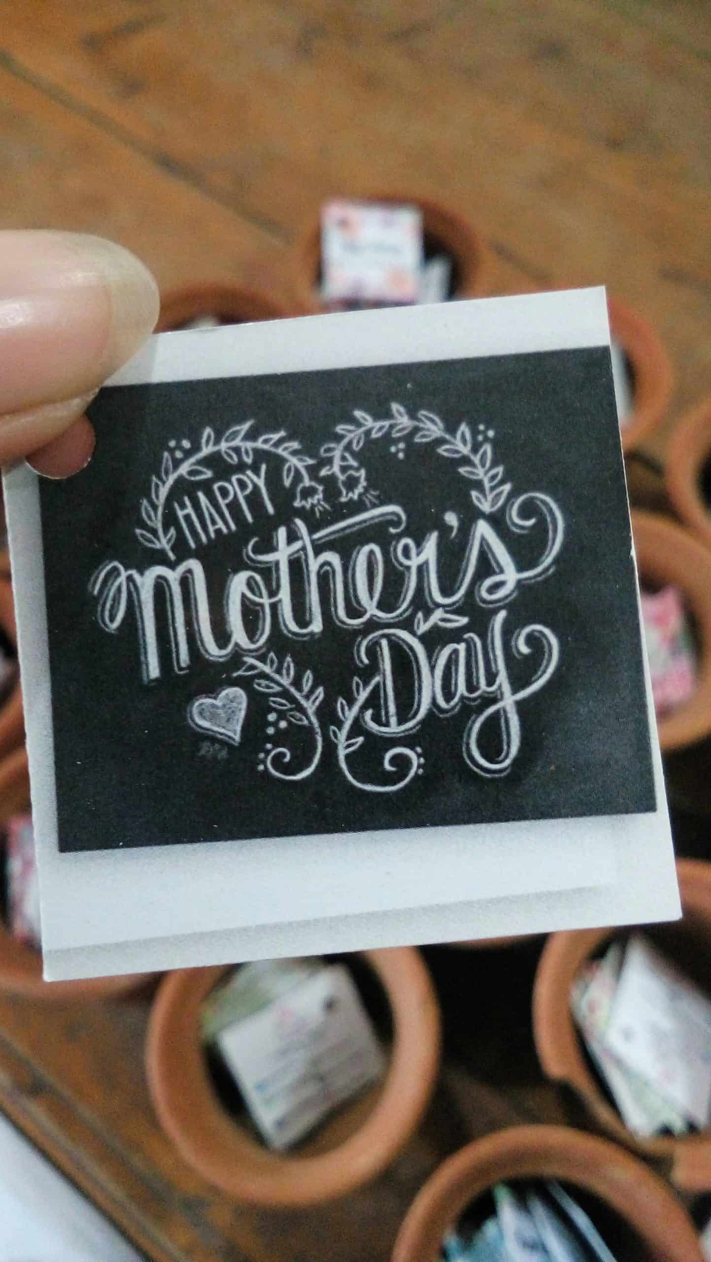 hand holding a happy mother’s day card with decorative lettering and small clay pots in the background