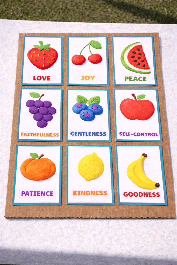 fruit of the spirit cards glued onto cardboard in a 3×3 grid showing love, joy, peace, faithfulness, gentleness, self-control, patience, kindness, and goodness on a white table outdoors