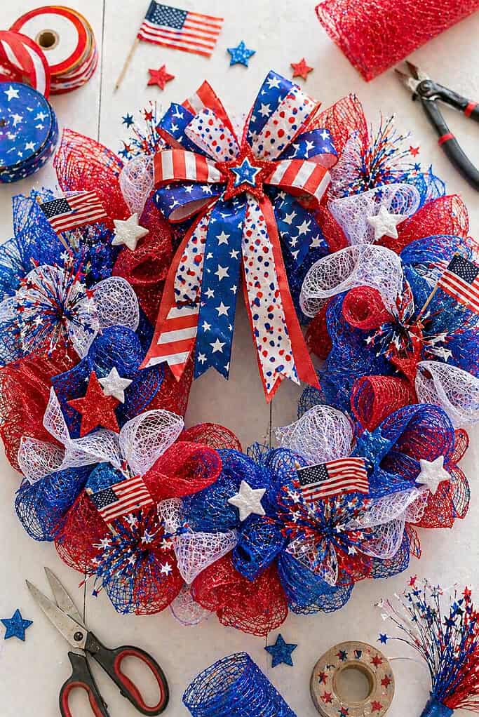 A full, circular wreath made from a wire or foam base, generously wrapped in red, white, and blue mesh ribbon.