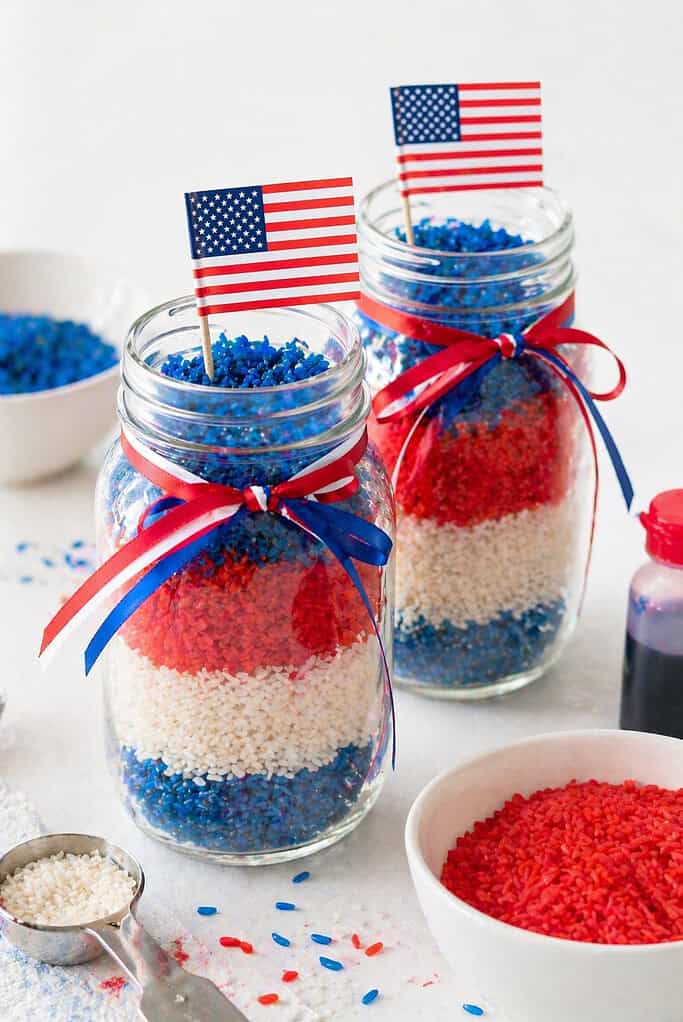 two clear glass mason jars or simple glass vases filled with neatly layered rice in patriotic colors—vivid red, bright white (natural rice), and deep blue.