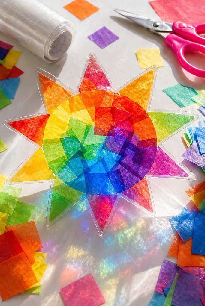 a finished suncatcher made from layered squares of translucent tissue paper in vibrant rainbow colors—reds, oranges, yellows, greens, blues, and purples