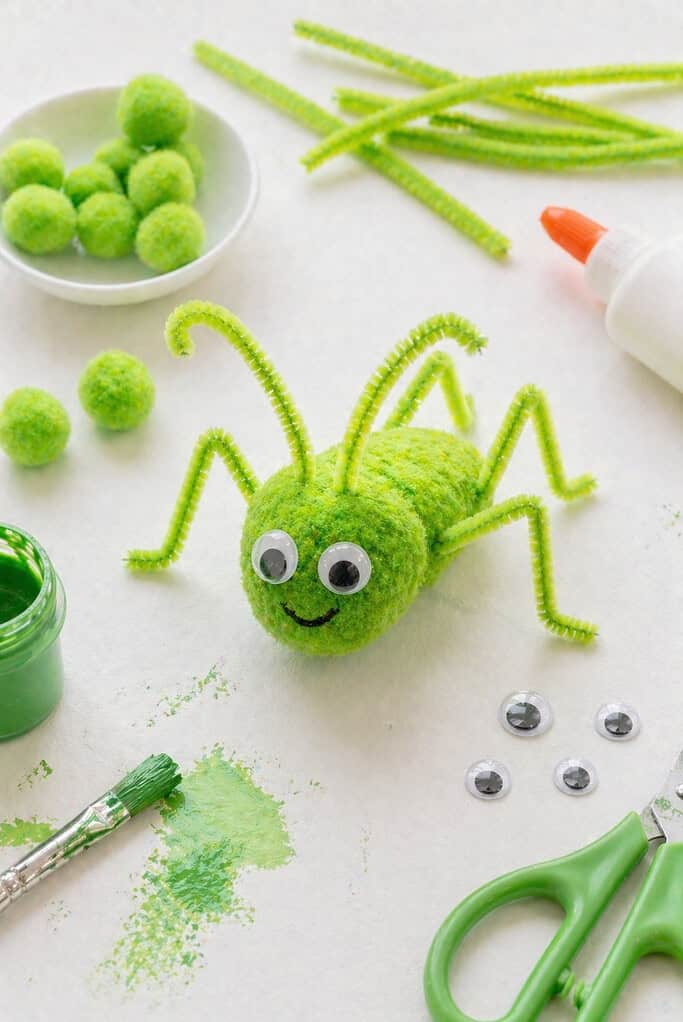 A small green pom pom (or a softly painted cotton ball) forms the cricket’s round body, with a slightly fluffy, textured surface.