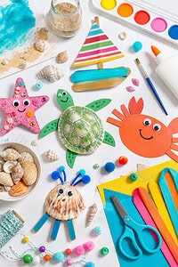 an assortment of beach-inspired kids’ craft supplies such as seashells, sand dollars, colored paper, googly eyes, popsicle sticks, child-safe scissors, paintbrushes, watercolor paints, markers, glue, twine, beads, pom-poms, and small jars of sand.