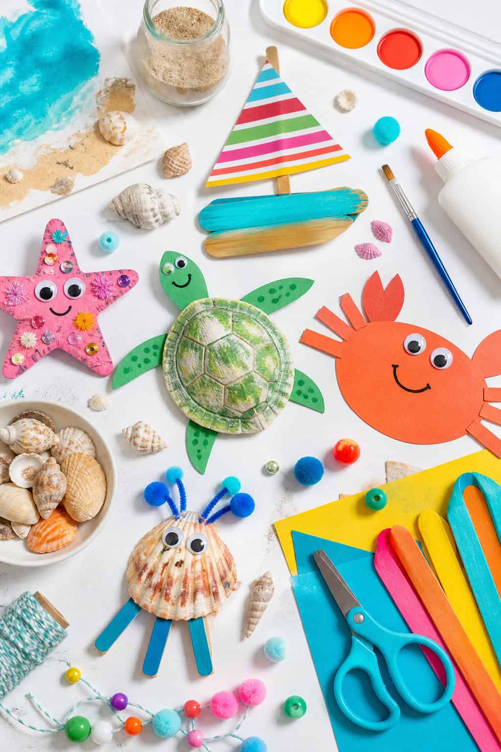 an assortment of beach-inspired kids’ craft supplies such as seashells, sand dollars, colored paper, googly eyes, popsicle sticks, child-safe scissors, paintbrushes, watercolor paints, markers, glue, twine, beads, pom-poms, and small jars of sand.