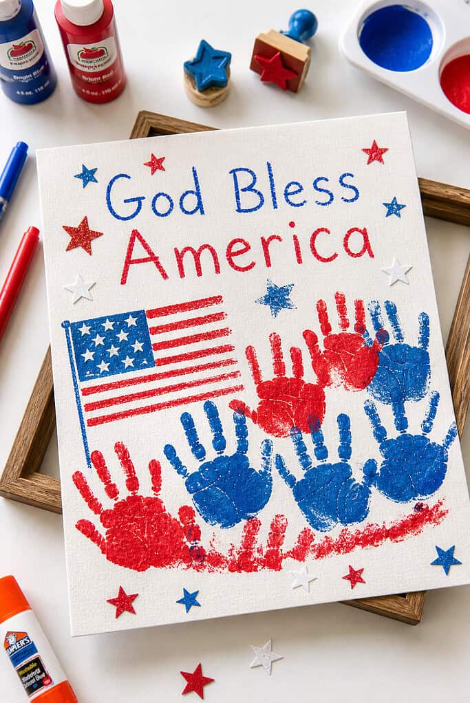A piece of textured canvas paper or sturdy white cardstock featuring the phrase “God Bless America” written across the top in soft, imperfect, childlike lettering.
