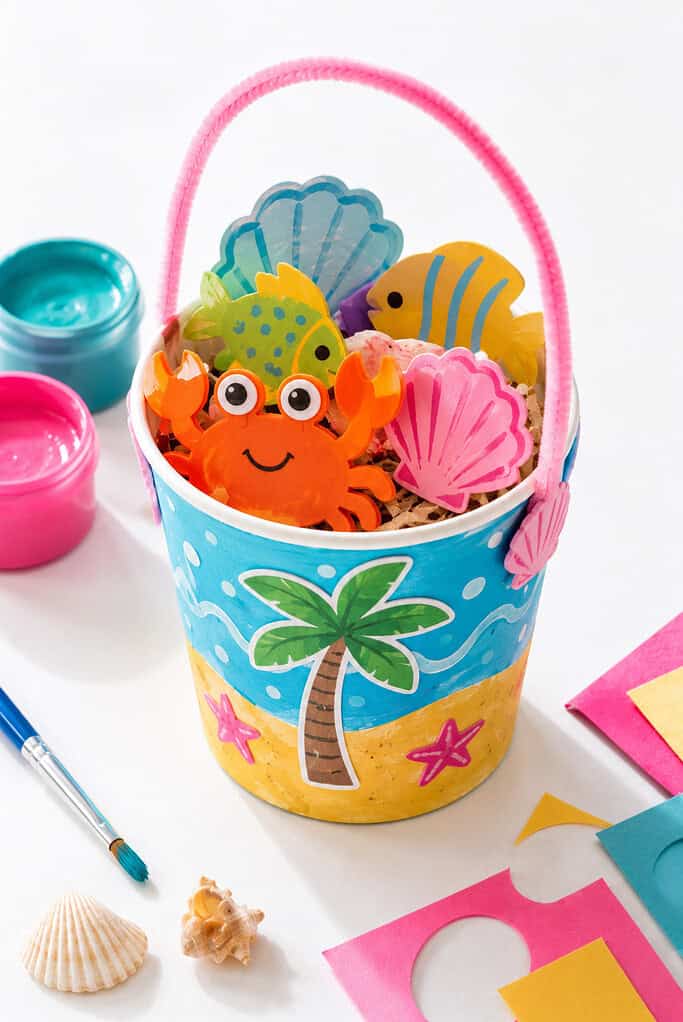 A cute handmade beach bucket craft made from a paper cup or cardboard tube decorated with bright paint, stickers, and simple beach-themed designs.