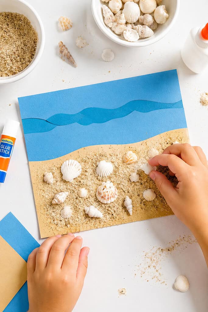 a simple handmade preschool beach collage created from cardstock: a strip of textured blue paper representing ocean water across the top and a strip of golden sand below