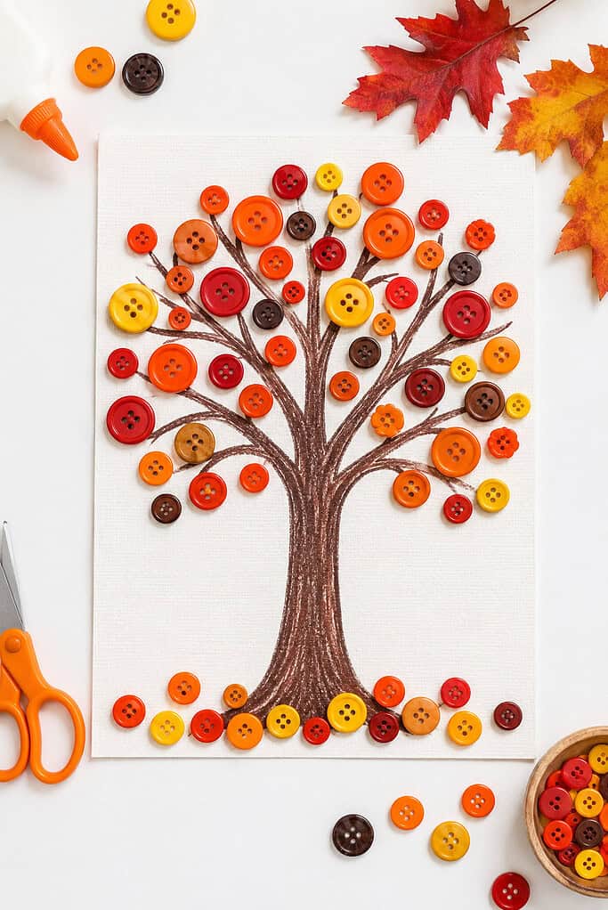 a handmade autumn tree artwork on textured cardstock or watercolor paper, featuring a simple brown tree trunk with branches filled with colorful buttons in rich fall shades