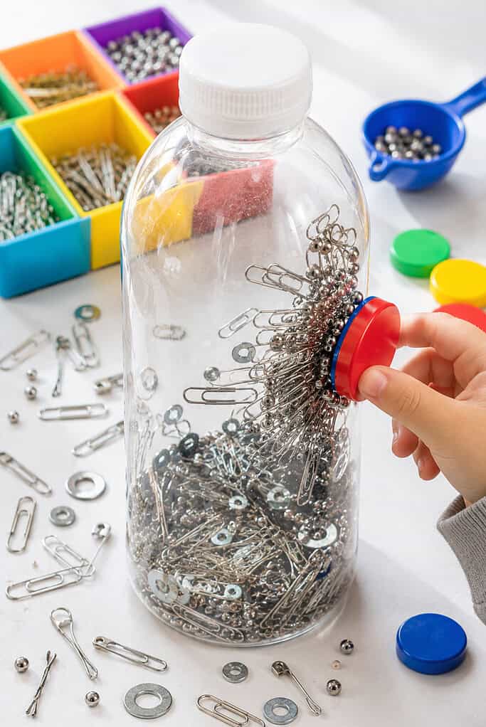 one clear plastic sensory bottle filled only with small metal treasures such as paperclips, washers, tiny metal beads, safety pins, and bobby pins inside the empty transparent bottle.