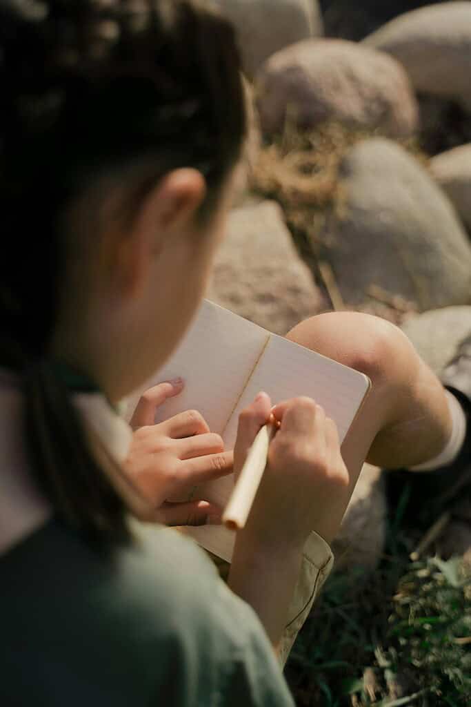A girl writes in a notebook while sitting on rocks outdoors.