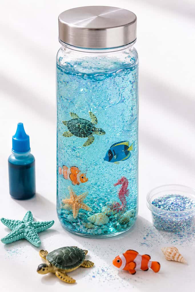 A clear sensory bottle filled with water tinted with soft blue food coloring, tiny plastic sea creatures, floating glitter, and subtle bubbles suspended throughout the bottle.