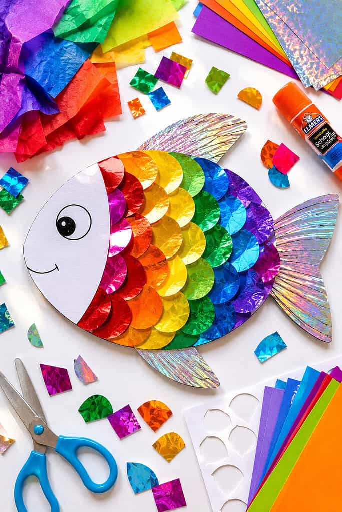 A large fish shape made from a light-colored cardstock, filled with overlapping metallic and iridescent paper scales in bright rainbow colors.