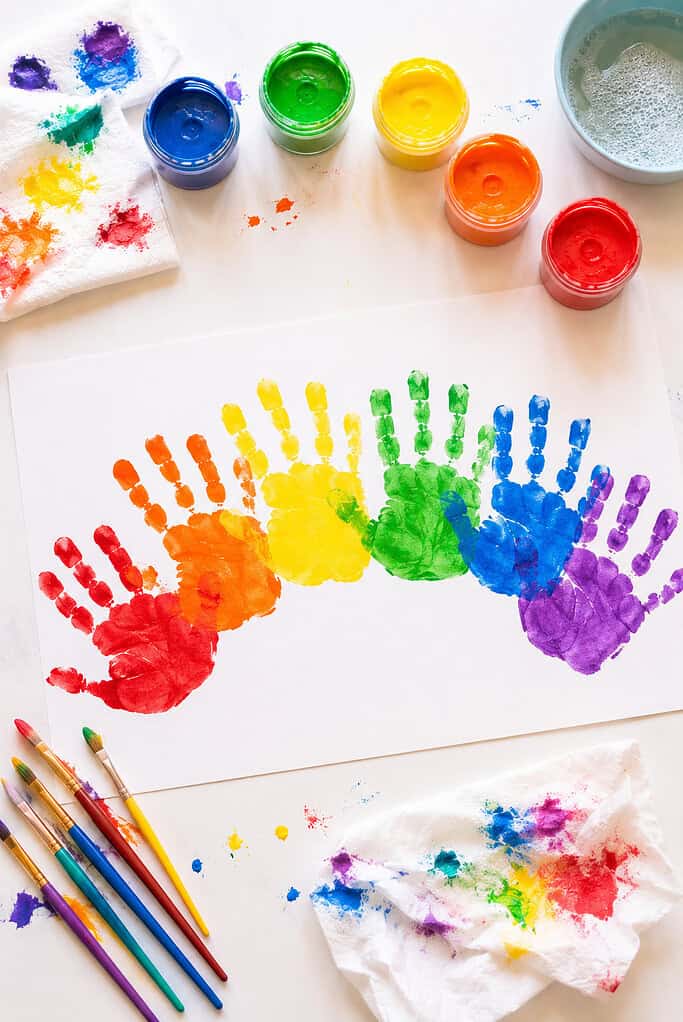child handprints painted in rainbow colors arranged in a curved rainbow arc across white cardstock